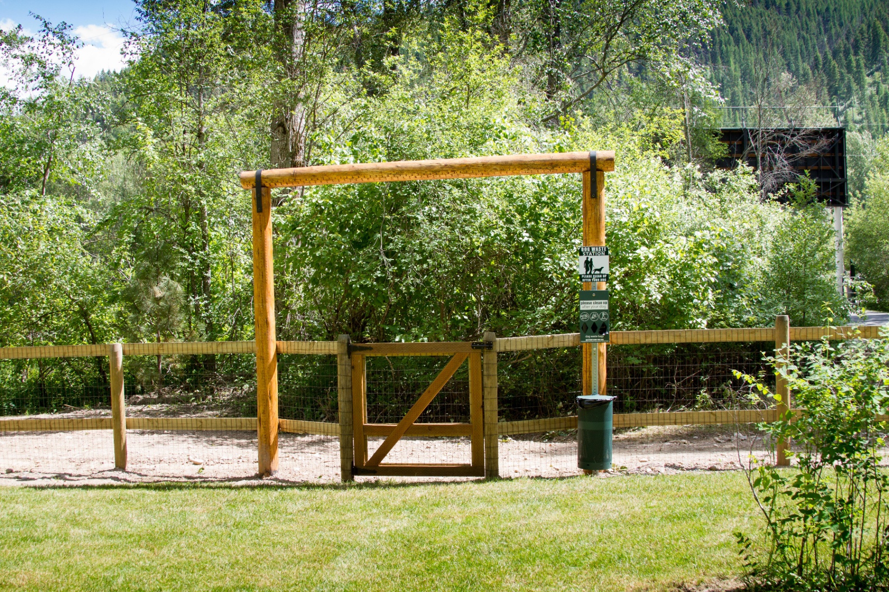 Fenced off-leash dog park entrance with wooden gate and waste station surrounded by green trees at Campground St. Regis