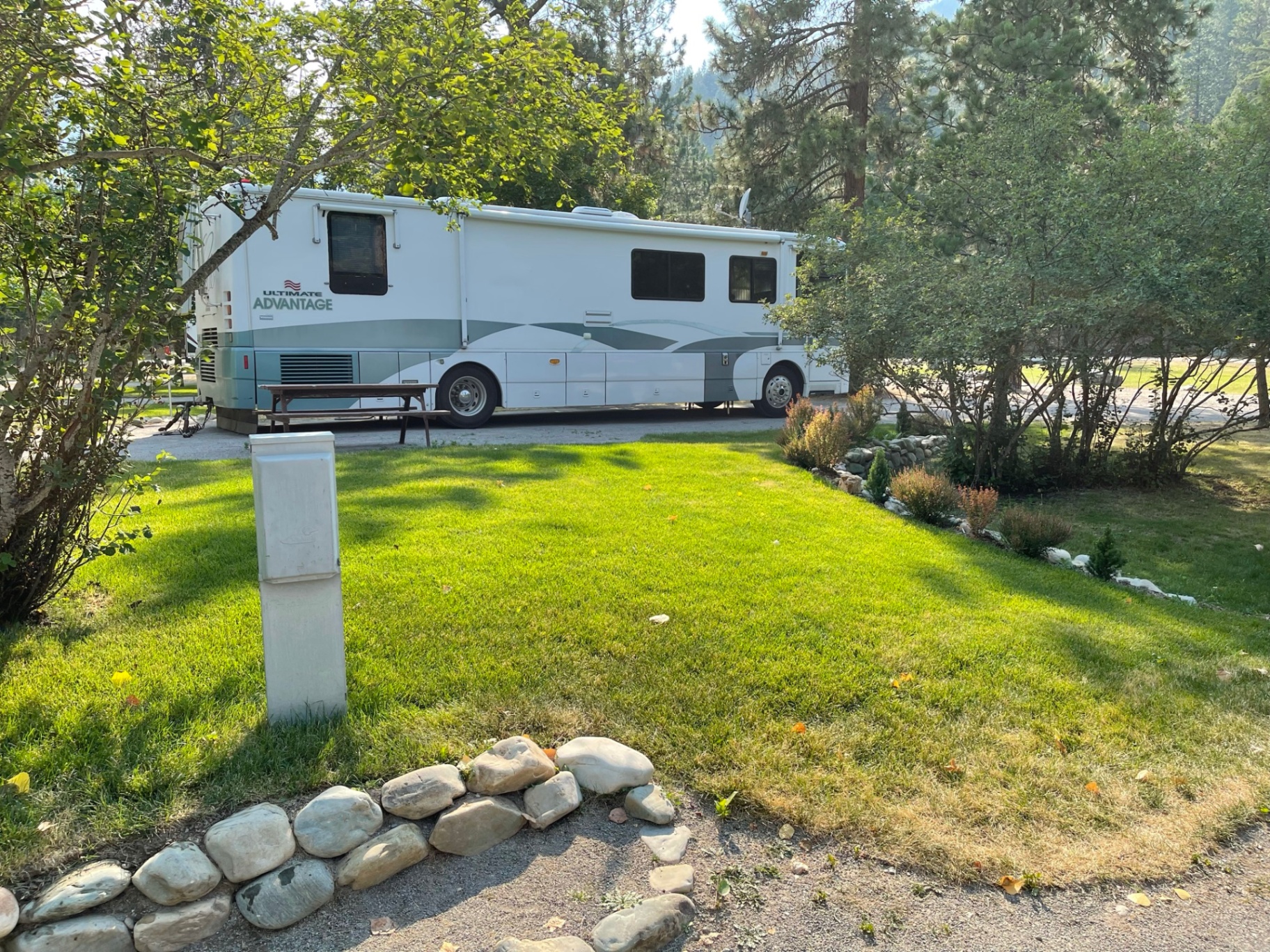 Grassy pull-through RV site at Campground St. Regis with a 50/30/20-amp breaker-protected pedestal in the foreground and a large motorhome parked among pines