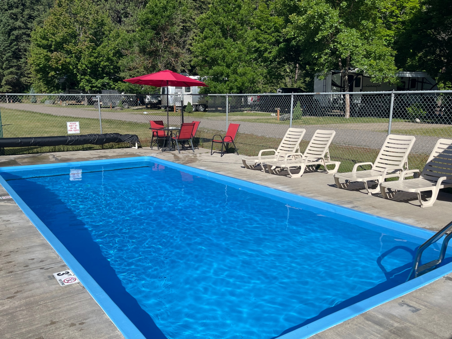 Heated outdoor swimming pool with bright blue water, red umbrella and chairs, white lounge chairs, and RV sites in the background at Campground St. Regis