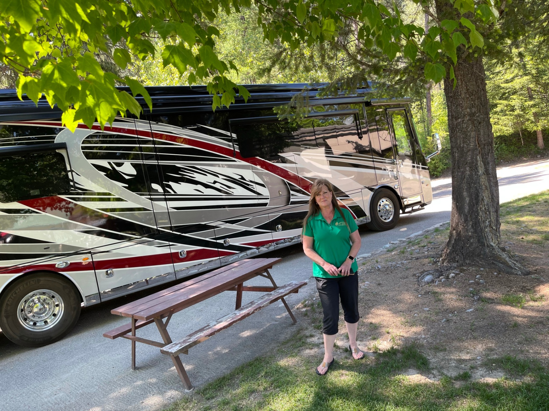 Camp host in a green polo standing beside a large luxury Class A motorcoach under shady maples