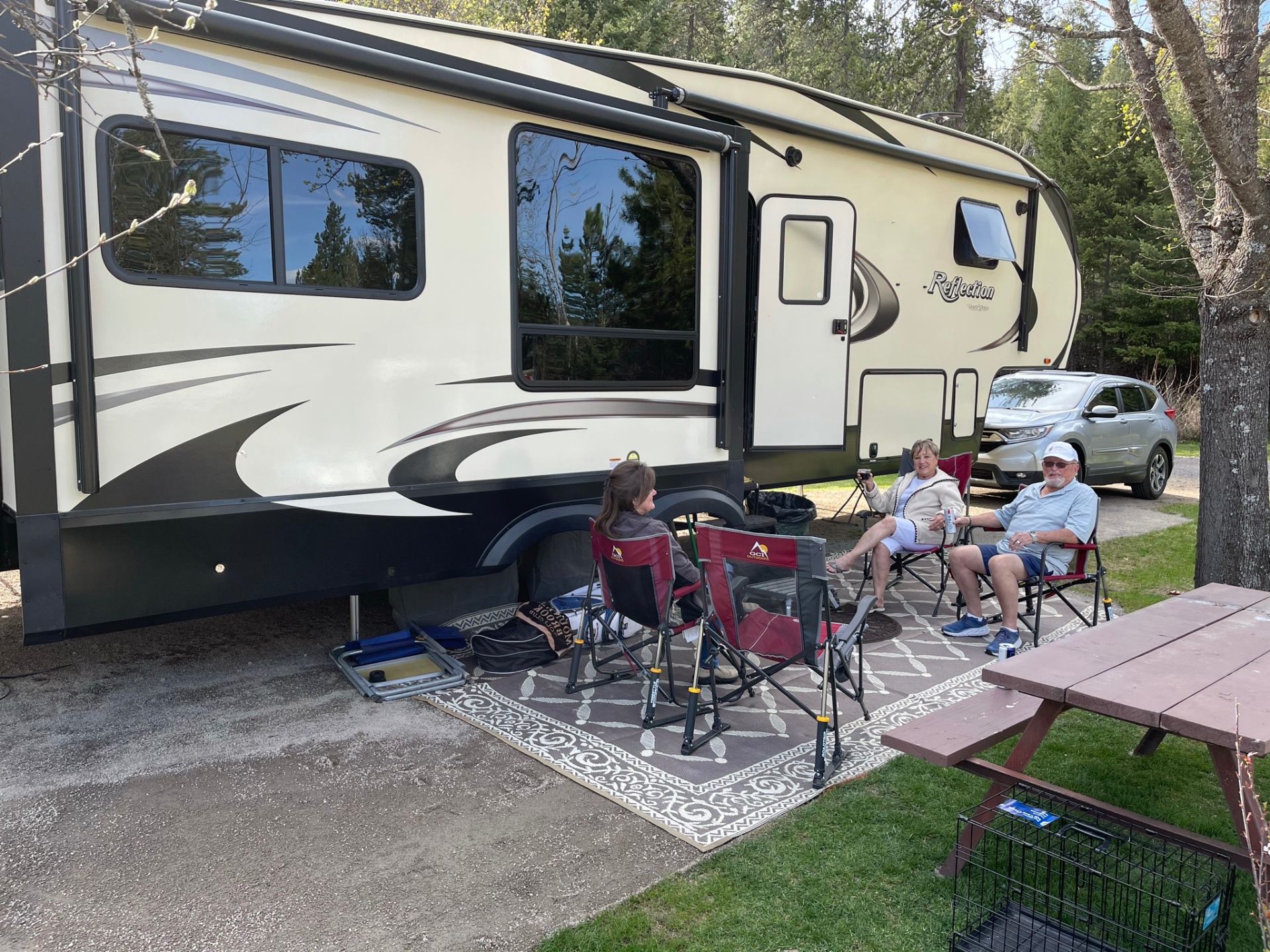 Fifth-wheel RV parked on a spacious wooded site with guests enjoying the outdoors